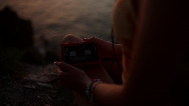 Young woman holds a red portable cassette player beside the ocean during warm sunset light, dusk final glow, closeup on analog device