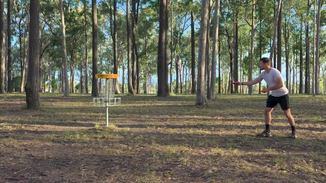 Caucasian man throwing a disc and hitting the net playing disc golf in a public park under trees on a sunny day in Logan, Brisbane, Queensland, Australia.