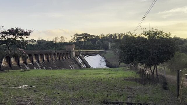 Zoom in toward a hydroelectric dam and spillway surrounded by vegetation at sunset in the rural landscape of Brotas, Brazil.