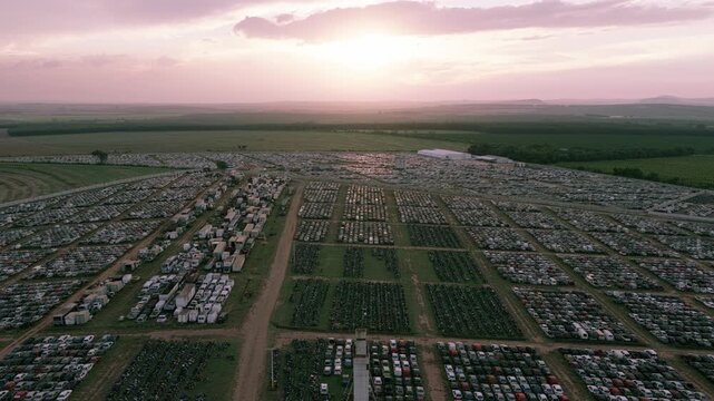 Aerial pan left over thousands of stored vehicles arranged in geometric sections across a huge logistics facility during sunset.