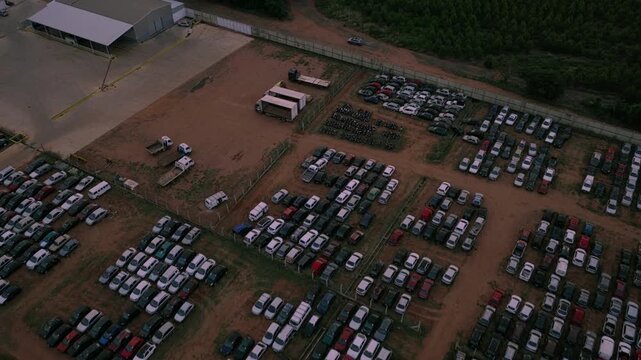 Descending aerial zoom in transitioning toward a zenithal perspective over organized rows of cars inside a large automotive storage facility.