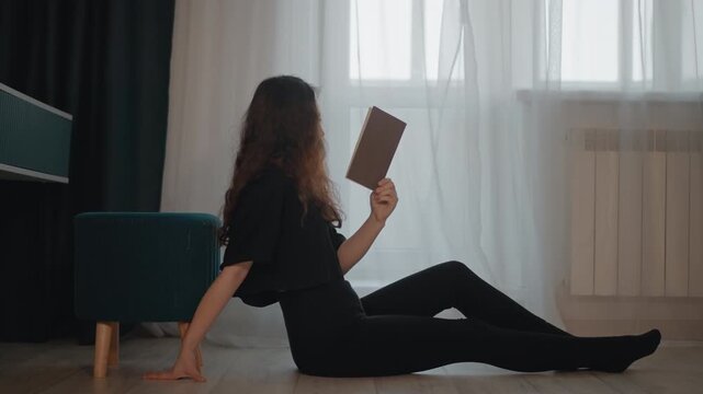 Young woman sits on hardwood floor beside tall window, reading book with relaxed posture, extended legs, and crossed ankles in calm minimalist living room filled with soft natural light and quiet