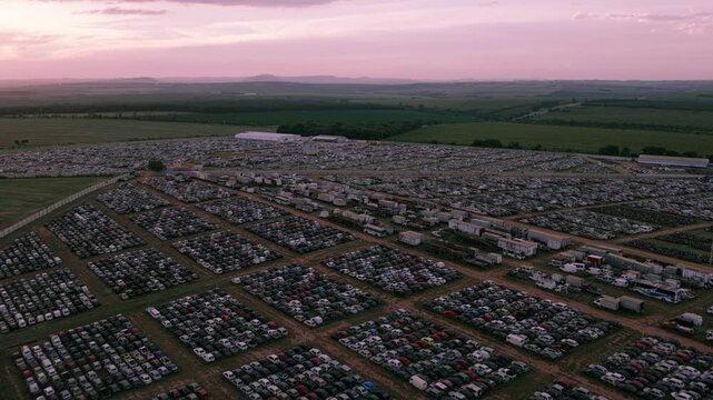 Aerial zoom in toward an enormous vehicle storage facility with thousands of cars organized in rows across a rural landscape.