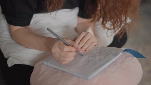 Overhead close framing shows two people seated with open notebook resting on pale cushion; visible action is right hand writing with pen while left hand steadies page, indicating active note-taking