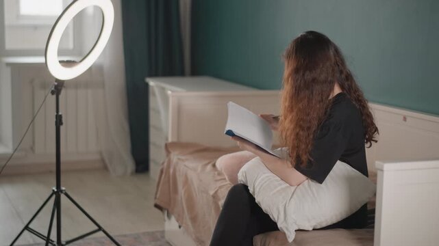 Young woman seated on bed reading and flipping papers in softly lit bedroom, with slight lean and hand movement suggesting focus, home study, quiet organization, and relaxed domestic routine