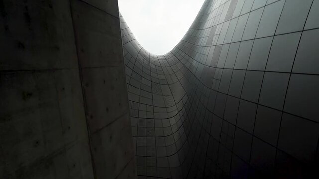 Curved metal panels rise between dark concrete walls as the camera tilts up toward a bright sky.