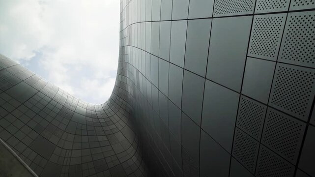 Camera tilts up along a perforated metal grid facade that curves inward toward a bright cloudy sky.