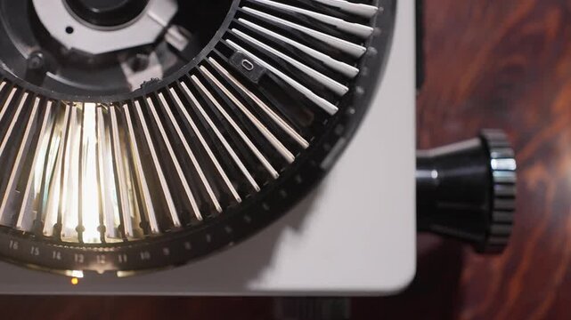 Vintage slide projector carousel glows as mounted photographic slides pass the lamp, overhead backdrop as light illimunates inside machine