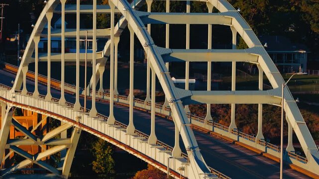 White arch bridge and roadway glow in warm autumn light among trees in Ozark, Arkansas, aerial telephoto tracking left with sunset glow