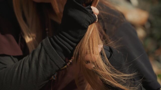 Tight indoor crop shows hands, hair, and beaded necklace as person runs fingers through straight blonde strands, creating gentle movement, soft glow, and relaxed beauty mood, with hair, necklace
