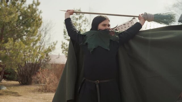 Single woman in layered dark cloak performs controlled staff movements in outdoor garden, with pointed stance, overhead lift, sweeping swing, diagonal reach, flowing fabric, soft daylight, and tense