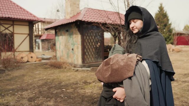 Woman in hooded cloak bends protectively beside small child holding brown hat while moving through rustic yard with gazebo, fence, bare trees, muted grass, and soft overcast light, with woman, child