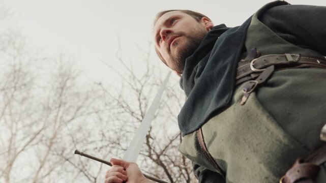 Bearded adult man grips sword with both hands, holding raised defensive pose beneath bare winter trees; focused upward gaze, tense shoulders, and rugged clothing suggest readiness for combat or