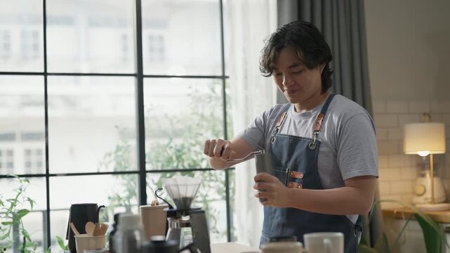 Young man in denim apron pouring freshly brewed coffee from carafe into cup, close-up action shot with soft window light, artisan coffee brewing process and morning ritual concept at home.