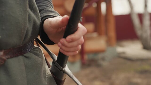 Close view of person in green wool tunic tightening and loosening aged leather belt while gripping polished metal sword hilt at waist, showing readiness, motion, and detailed costume texture in soft