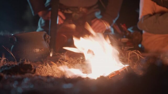 Close low view of lively campfire at night with flickering flames, rising sparks, glowing embers, smoke, and partial hands and legs of seated people gathering nearby for warmth and relaxed outdoor
