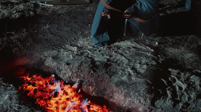 Bound soldier kneels on rough ground in chainmail coif and dark cloak, head turned with wary tension while ember light and cold moonlight shape worn armor, rope restraint, helmet, dirt, and