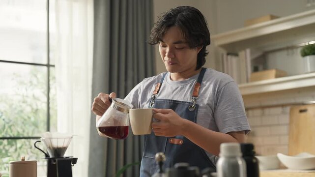 Young man in denim apron pouring freshly brewed coffee from carafe into cup, close-up action shot with soft window light, artisan coffee brewing process and morning ritual concept at home.