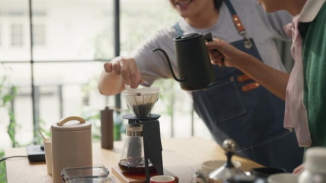 Man in denim apron smiling while brewing pour-over coffee, teaching friend at home kitchen counter, coffee beans and drip equipment on table, weekend hobby and hospitality concept, warm morning light.
