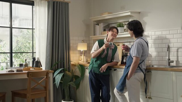 Two men chatting and laughing while drinking coffee in cozy home kitchen, one wearing denim apron, morning routine and casual conversation, warm domestic lifestyle concept with natural light.