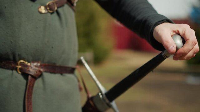 Close-up of person in green medieval-style tunic and leather belt gripping sword hilt near pommel and crossguard while slowly revealing blade from scabbard at waist in soft daylight