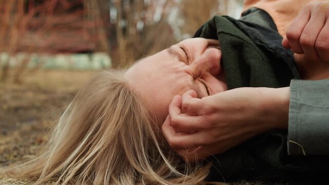 Close-up of young person lying on brown grass and soil outdoors, face tightly scrunched, eyes closed, hands near face, with tousled hair, tilted head, and natural daylight creating earthy texture
