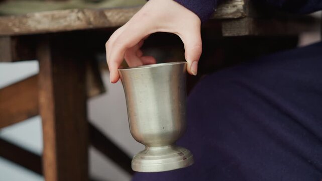 Adult hand lifts small metal goblet from rustic wooden table edge, fingers curved around rim and base while gentle rotation reveals careful grasp, open space below, and worn wood texture