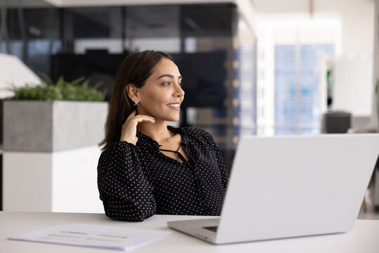 Young Arabian businesswoman sitting at office desk with papers and open laptop, looks aside with smile, taking thoughtful break, enjoying moment of reflection during busy workday. Career perspectives