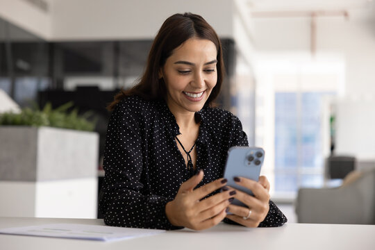 Cheerful woman sits at workplace desk in modern office, smile, looking at smartphone screen, reading personal message with good news, review work results, feel satisfied. Apps, tech usage in business