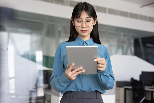 Focused Asian businesswoman standing in contemporary office, using tablet, makes business-related tasks, reviewing project reports, checking analytics data or sales performance, research market trends