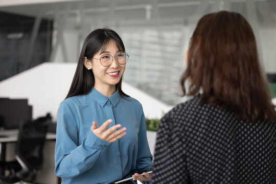 Young Asian professional woman having friendly discussion, informal conversation with female colleague standing in modern coworking space. Business communication, collaboration, workplace interaction