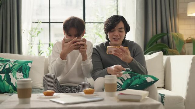 Asian gay couple laughing together while playing mobile game on smartphone, relaxing on sofa at home, joyful bonding moment, LGBTQ relationship and technology lifestyle concept, natural indoor light.