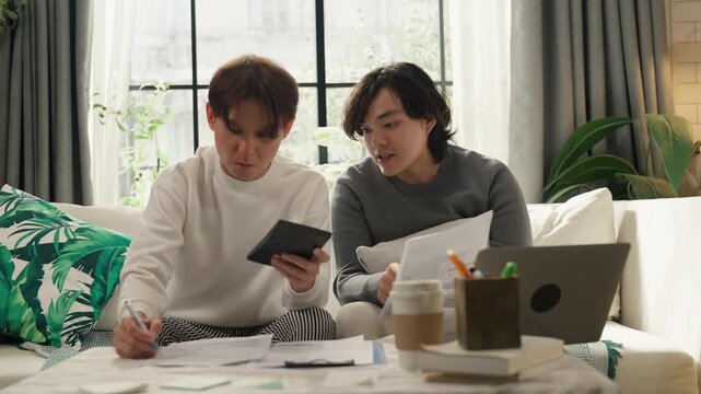 Two men sitting on sofa at home reviewing bills and calculating household budget together, holding calculator and paperwork, personal finance and expense management concept, cozy living room with lapt