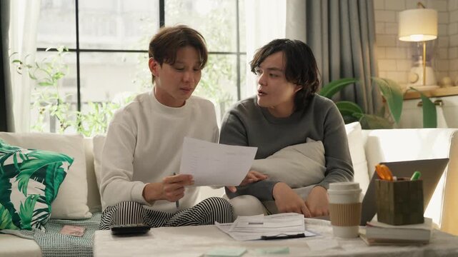 Two men sitting on sofa at home reviewing bills and calculating household budget together, holding calculator and paperwork, personal finance and expense management concept, cozy living room with lapt