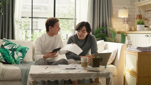 Two men sitting on sofa at home reviewing bills and calculating household budget together, holding calculator and paperwork, personal finance and expense management concept, cozy living room 