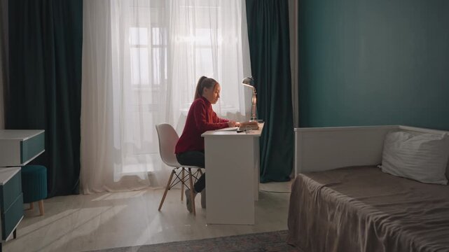 Young woman in red top is seated at white desk engaged. Room features teal walls with dark green drapes and natural light. Metallic lamp illuminates workspace as woman interacts