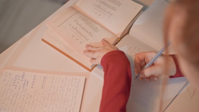 Young woman with long hair writes. Her left hand rests near opened book. Notebook page to bottom left shows dense handwriting including dates like 14 10 25 and language learning