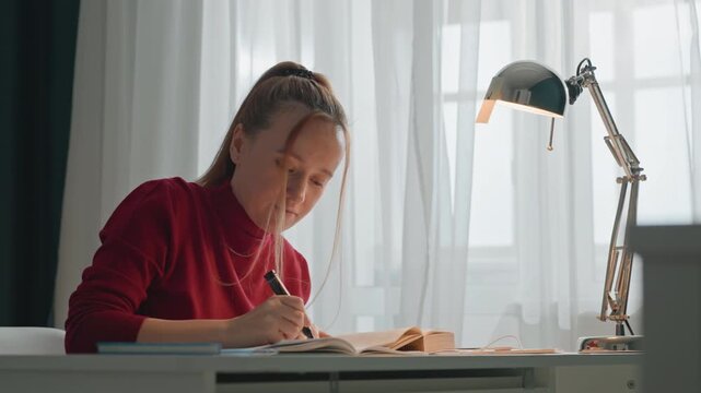 Young woman in red sweater studies at desk writing. Woman adjusts lamp shade while interacting with study materials. Scene is well lit by natural light through sheer curtains