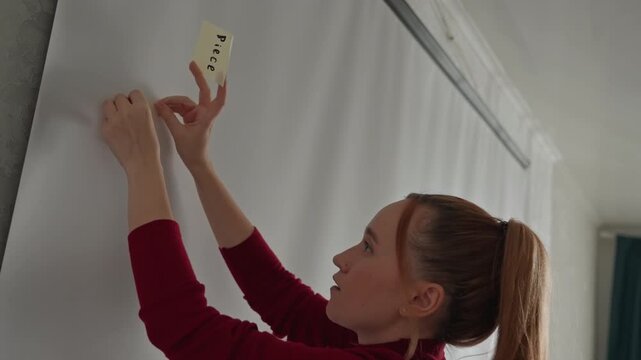 Young woman in red turtleneck examines and places sticky. Woman adjusts note positions with both hands focusing intently. Bright room features sheer curtains dark green drapes
