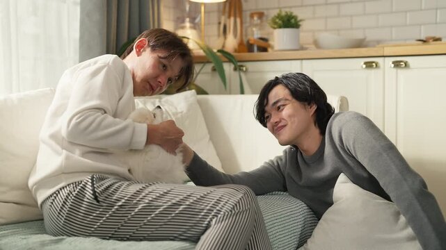 Two smiling friends feeding treat to fluffy Pomeranian puppy on sofa at home, playful moment with dog, pet ownership and companionship concept, cozy living room, natural indoor light.