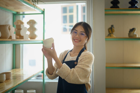 Smiling Asian Woman Artisan Holding Ceramic Cup in Her Small Pottery Studio Shop