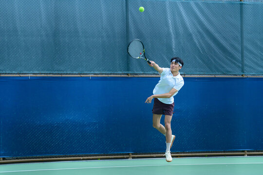 Young Asian Male Tennis Player Hitting a Powerful Serve on Outdoor Court
