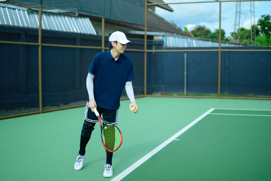 Asian Male Tennis Player Standing on Outdoor Court Holding Racket and Ball with Knee Brace