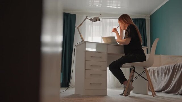 Young woman with long hair sits at white desk. Room features teal walls natural light from windows covered. Woman remains focused at desk under consistent lighting