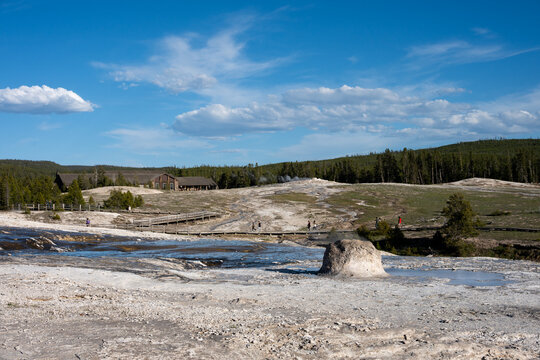 Beehive Geyser erupting in early morning, tourists on boardwalk with visitor center in background, Upper Geyser Basin, summer vacation exploring Yellowstone National Park
