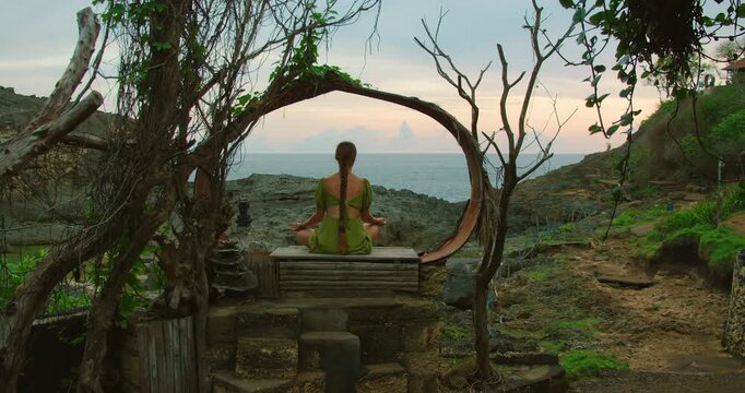 Woman meditates in lotus pose within circular wooden frame overlooking ocean in Nusa Penida Bali
