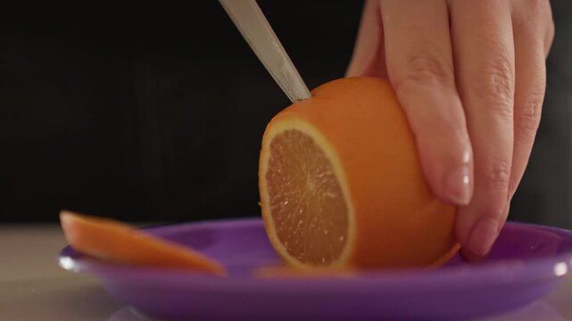 Close up of hands slicing orange on purple plate. Orange is cut into thin segments revealing juicy interior texture. Dark background emphasizes cutting action and fruit details
