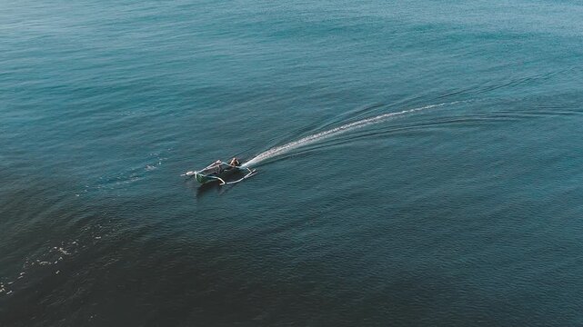 Drone aerial footage of a small traditional outrigger fishing boat with a curved wake trail sailing on vast deep blue ocean. Calm sea with contrast of light and dark water. Minimalist seascape and fis
