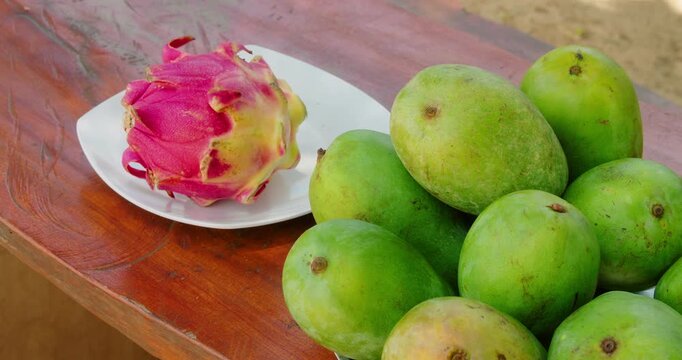 Dragon fruit and green mangoes displayed on wooden table in tropical setting