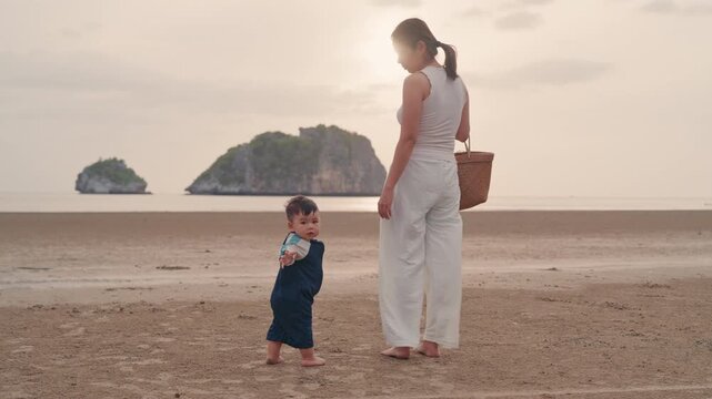 Asian mother holding hands with toddler son walking on sandy beach together with sunset.